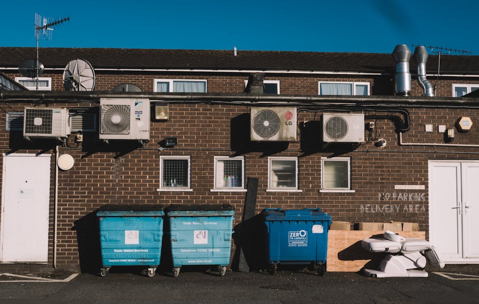 A row of three large wheelie bins, two blue and one darker blue, are positioned on the pavement in front of a brick building with small windows and an external wall-mounted air conditioning system. The building's exterior features multiple air conditioning units and ducts mounted on the wall, indicative of climate control infrastructure. To the right of the bins, there are two white plastic furniture pieces, possibly chairs or stacked items, placed on the ground near a wooden pallet. The pavement is clean and the scene is evenly lit by daylight, with a clear blue sky overhead. This setting illustrates a typical outdoor space for waste disposal and storage, common in residential or commercial properties, aligning with house removals and moving services by Man with Van East Bedfont, especially when coordinating the disposal of bulky waste or preparing for a home relocation.