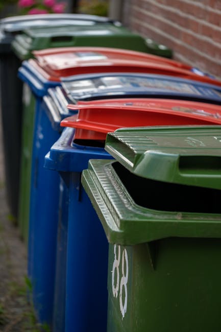 A row of four large wheelie bins positioned outdoors along a pavement beside a brick wall. The bins are arranged in a line, with the closest being green, followed by blue, red, and another green bin visible in the background. Each bin has a matching lid, with some lids slightly open or closed, revealing the internal capacity for waste collection. The bins are used for household waste disposal and are part of a home relocation or waste management process. The scene is captured in natural daylight, highlighting the textures and colors of the plastic bins, which are commonly used by moving or removal services such as Man with Van East Bedfont for efficient waste and bulky item disposal during relocations.