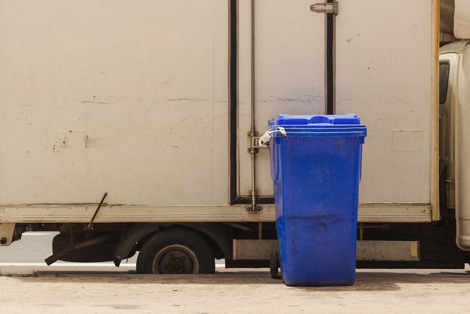 A row of three large wheelie bins, two blue and one darker blue, are positioned on the pavement in front of a brick building with small windows and an external wall-mounted air conditioning system. The building's exterior features multiple air conditioning units and ducts mounted on the wall, indicative of climate control infrastructure. To the right of the bins, there are two white plastic furniture pieces, possibly chairs or stacked items, placed on the ground near a wooden pallet. The pavement is clean and the scene is evenly lit by daylight, with a clear blue sky overhead. This setting illustrates a typical outdoor space for waste disposal and storage, common in residential or commercial properties, aligning with house removals and moving services by Man with Van East Bedfont, especially when coordinating the disposal of bulky waste or preparing for a home relocation.
