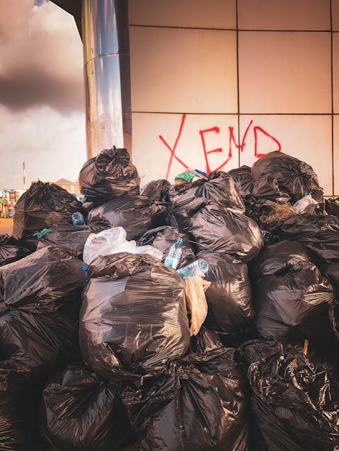 A mound of tightly packed black rubbish bags filled with household waste is stacked against a modern exterior wall of a building with large, shiny metal panels. Some bags are partially torn, revealing plastic bottles and miscellaneous trash inside. The bags are placed outdoors on pavement, with a clear sky and cloud formation visible in the background. Red graffiti spelling 'XEND' is spray-painted on the wall behind the waste. This scene illustrates improper waste disposal, relevant to waste rules and council regulations discussed in the context of avoiding fines in East Bedfont. Such waste collection issues can impact home relocation or moving services like those provided by Man with Van East Bedfont, especially when clearing properties before a move.