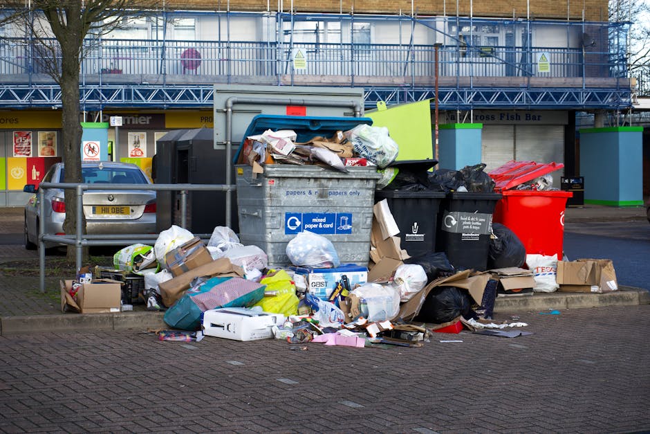 A large outdoor waste disposal area in East Bedfont with multiple overflowing recycling and general waste bins. The bins are situated on a paved pavement next to a metal railing, with miscellaneous rubbish spilling onto the ground, including cardboard boxes, paper, plastic bags, and packaging materials. Some cardboard boxes are flattened or partially collapsed, and several black and black-and-white plastic bin bags are piled around the bins. The background features a building with scaffolding, a shop with a yellow sign, and parked cars, one of which has a partially visible license plate. The scene illustrates the importance of proper waste management in residential and commercial areas, relevant to house removals and moving services provided by Man with Van East Bedfont, especially in relation to complying with council waste rules during home relocation processes.