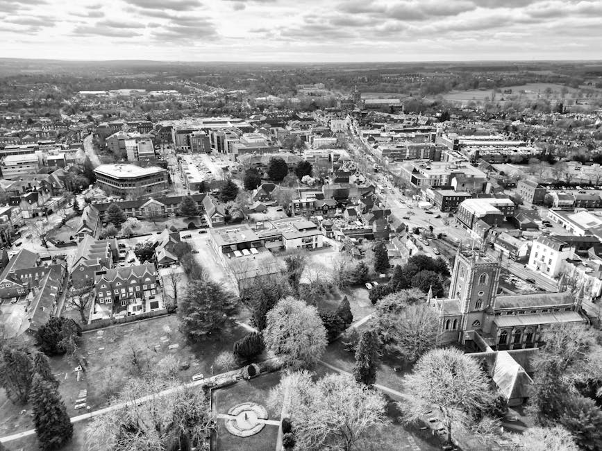 Aerial black-and-white photograph of a townscape featuring a mix of residential and commercial buildings, with a prominent church with tall steeple near the foreground. Surrounding the church are trees, some without leaves, and well-maintained gardens. Streets and pathways are visible, with vehicles and pedestrians in the distance. The scene includes a park area with a circular feature, likely a fountain or sculpture, and the overall environment suggests a typical urban area with a combination of older and modern structures, captured during daytime under partly cloudy skies. This setting supports the context of home relocation and moving logistics by illustrating a typical East Bedfont locality scene with urban infrastructure and community spaces, relevant to house removals and furniture transport services.