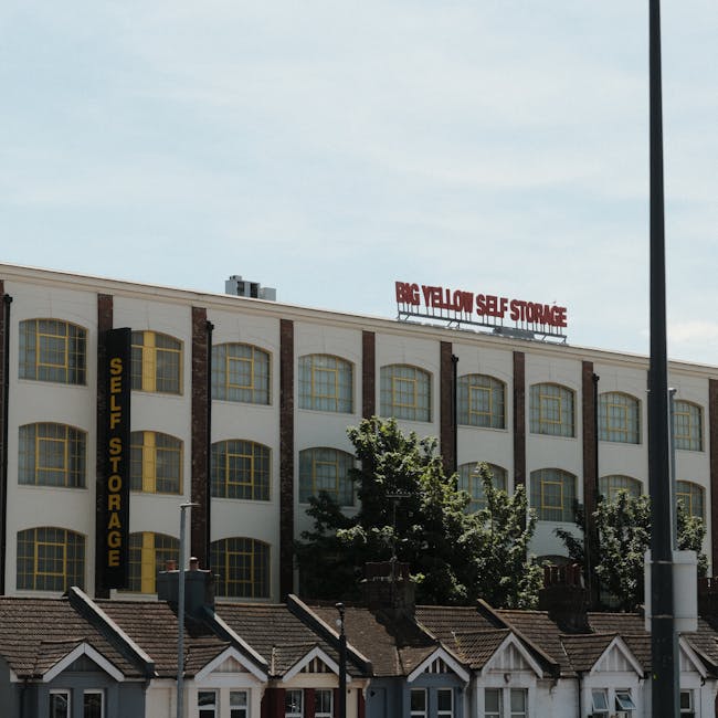 A large multi-storey white building with a brick accent façade housing Big Yellow Self Storage, featuring multiple arched windows with yellow window frames. A vertical yellow sign on the side reads 'SELF STORAGE' in black lettering. At the top of the building, a red and white sign displays 'BIG YELLOW SELF STORAGE'. In front of the building, there are small residential houses with pitched roofs and chimneys, partially obscured by green trees and bushes. The sky above is clear with some light cloud cover, and a tall street lamp is visible on the right side of the image. The scene appears to be taken during daytime, highlighting a commercial property used for storage services, relevant to home relocation or furniture transport involved in moving and packing processes.