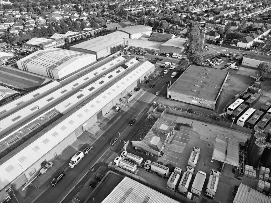 Aerial black-and-white photograph of a townscape featuring a mix of residential and commercial buildings, with a prominent church with tall steeple near the foreground. Surrounding the church are trees, some without leaves, and well-maintained gardens. Streets and pathways are visible, with vehicles and pedestrians in the distance. The scene includes a park area with a circular feature, likely a fountain or sculpture, and the overall environment suggests a typical urban area with a combination of older and modern structures, captured during daytime under partly cloudy skies. This setting supports the context of home relocation and moving logistics by illustrating a typical East Bedfont locality scene with urban infrastructure and community spaces, relevant to house removals and furniture transport services.