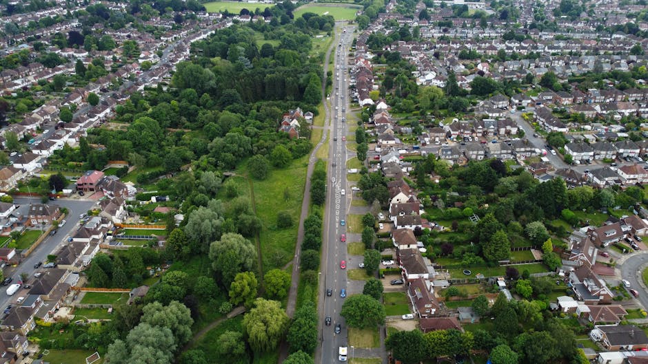 An aerial view of a suburban residential area showing rows of terraced houses with front gardens, parked cars lining the street, and a wide pavement. The scene captures a busy house relocation environ