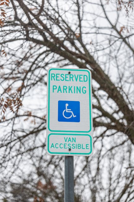 A close-up view of a reserved parking sign indicating accessible van parking, mounted on a metal pole outdoors against a background of tree branches with some remaining autumn leaves and an overcast sky. The sign features a blue wheelchair symbol and text reading 'RESERVED PARKING' at the top and 'VAN ACCESSIBLE' at the bottom, with the overall scene suggesting a location suitable for home relocation or furniture transport vehicles, as part of a moving process. The image emphasizes parking regulations relevant to residential moves, with natural lighting and clear signage support for moving logistics managed by Man with Van East Bedfont.