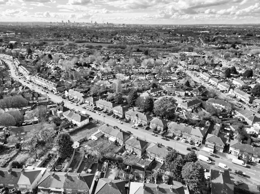 A black-and-white aerial photograph of a residential area in East Bedfont, showing a row of houses lined along a street with parked cars on both sides. The front yards are visible with some greenery and trees, and the rooftops are mostly pitched with tiles. In the foreground, rooftops and back gardens of individual homes are visible, while in the background, there is a wider view of the town extending to the horizon, including some larger buildings and open spaces. The scene captures the typical layout of suburban housing, with an emphasis on the street and surrounding residential infrastructure, relevant to the context of home relocation and furniture transport services as provided by Man with Van East Bedfont.
