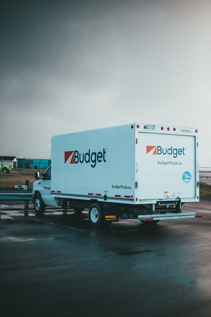 A white Budget moving truck is parked on a paved surface, with the company's logo and website displayed on the side and rear doors. The truck is positioned near an open area, possibly a parking lot or
