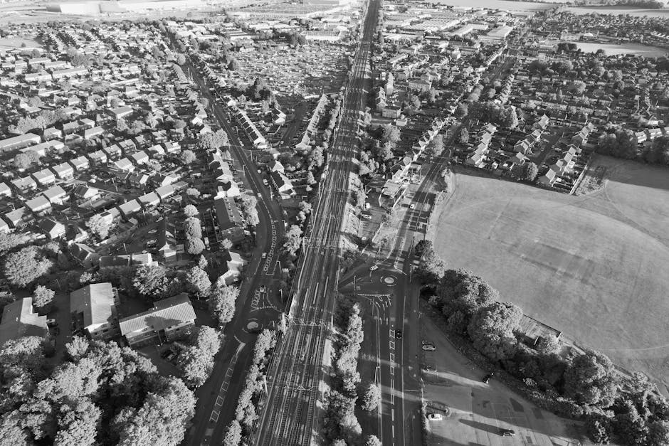 A high-angle black and white aerial photograph showing a residential neighbourhood with numerous houses and rows of trees along the streets. A set of railway tracks runs through the center, crossing a main road with multiple lanes, roundabouts, and traffic markings. To the right of the railway, there is a large open grassy field bordered by clusters of trees. Inside a property on the left side of the image, visible objects include stacked cardboard boxes, wrapped furniture covered in plastic or blankets, and a small moving trolley being used to load or unload household items. Near the entrance of the driveway, a van associated with Man with Van East Bedfont is partially visible, parked close to the property while the loading process occurs. The environment is well-lit with natural daylight, and the overall scene depicts a typical home relocation process involving furniture transport and the movement of packed items from the house to the vehicle, highlighting the logistics of residential moving services.