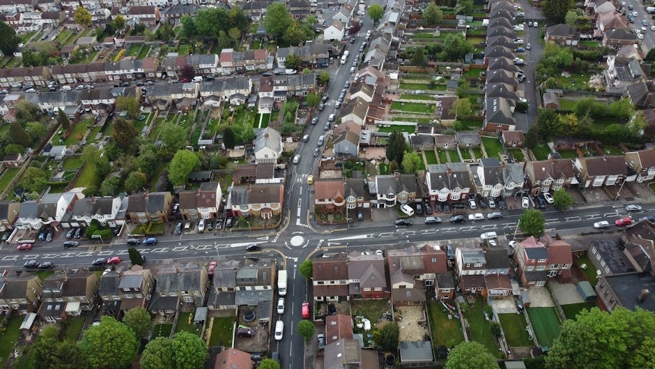 An aerial photograph of a residential neighbourhood showing a variety of houses, including semi-detached and terraced properties, arranged along a grid of streets. Several vehicles, such as cars and vans, are parked along the roadside and in driveways. In the foreground, a house with a backyard has visible packing materials like cardboard boxes and plastic wraps on the driveway, suggesting a home relocation process. A large van is parked outside the property, with the sliding door open and a person loading or unloading furniture and boxes via a trolley. The scene is well-lit with natural daylight, and trees and greenery are dispersed throughout the area. This visual context supports the services of Man with Van East Bedfont, specialising in removals, packing, and furniture transport in the area between Hatton Cross and East Bedfont.