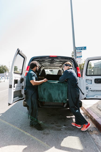 Two removal workers from Man with Van East Bedfont are loading a large, dark green upholstered item covered with protective fabric into the back of a white moving van, situated on a paved street with open van doors. The workers are dressed in dark uniforms and are carefully lifting and positioning the item during the home relocation process. Inside the van, visible wooden crates and cardboard boxes are organized for transportation. In the background, there are street signs, a lamppost, and parked vehicles, indicating a residential or commercial area. The scene is set during daylight hours with clear skies, providing natural lighting that highlights the team’s careful handling of the furniture and the detailed packing activities involved in furniture transport and packing and moving services.