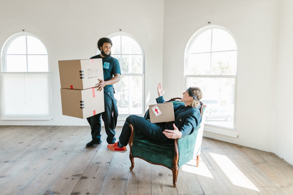 Two removal workers from Man with Van East Bedfont are loading a large, dark green upholstered item covered with protective fabric into the back of a white moving van, situated on a paved street with open van doors. The workers are dressed in dark uniforms and are carefully lifting and positioning the item during the home relocation process. Inside the van, visible wooden crates and cardboard boxes are organized for transportation. In the background, there are street signs, a lamppost, and parked vehicles, indicating a residential or commercial area. The scene is set during daylight hours with clear skies, providing natural lighting that highlights the team’s careful handling of the furniture and the detailed packing activities involved in furniture transport and packing and moving services.
