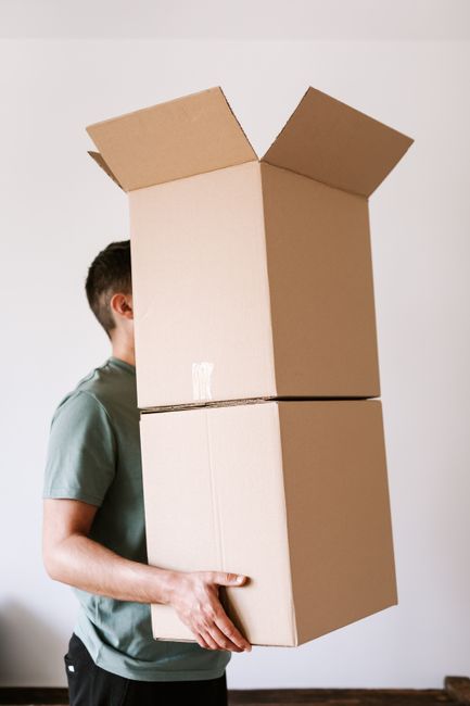 A person wearing a green t-shirt is holding a large cardboard box in each hand, with the boxes stacked one on top of the other. The top box's flaps are open and slightly bent outward, revealing its em
