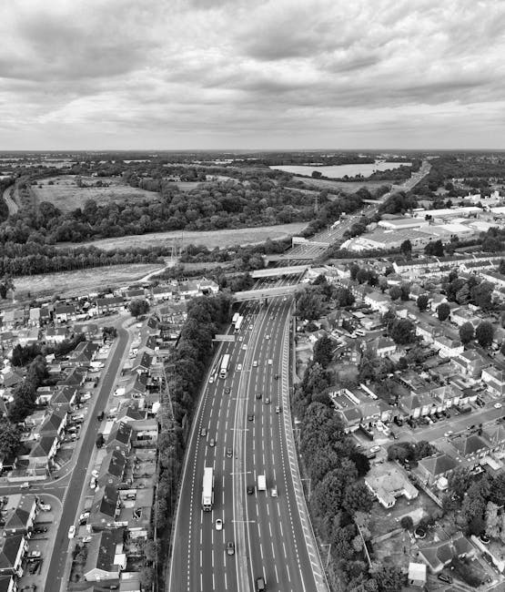 A black-and-white aerial photograph showing a residential area with houses, trees, and a multi-lane motorway running through the scene. The motorway is populated with various vehicles including cars and lorries, and is situated near a suburban neighbourhood with individual houses, gardens, and small streets. In the background, there are open fields and a river or large water body, with a cloudy sky overhead. This image exemplifies a typical urban and rural landscape, often involved in house removals and furniture transport logistics. The scene highlights the transitional environment encountered during home relocation processes, where transportation infrastructure such as busy roads and residential areas intersect with open countryside.