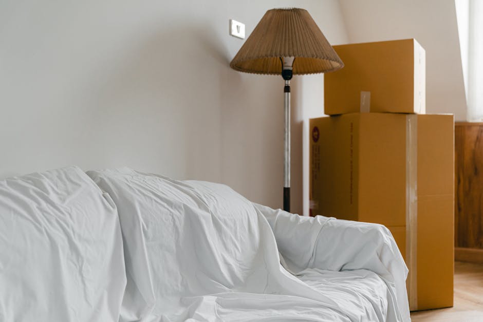Inside a living room during a home relocation, several brown cardboard boxes of varying sizes are stacked next to a wooden cabinet, ready for transport. A floor-standing lamp with a brown lampshade is positioned behind the boxes, near a plain white wall. The environment is well-lit with natural light coming from a nearby window. In the foreground, part of a white sofa covered with a protective cloth is visible, indicating ongoing packing or furniture moving preparations. The scene captures the loading or packing process typical of furniture transport and home removals, with objects arranged for easy handling by a team such as Man with Van East Bedfont, who specialise in removals and moving services.