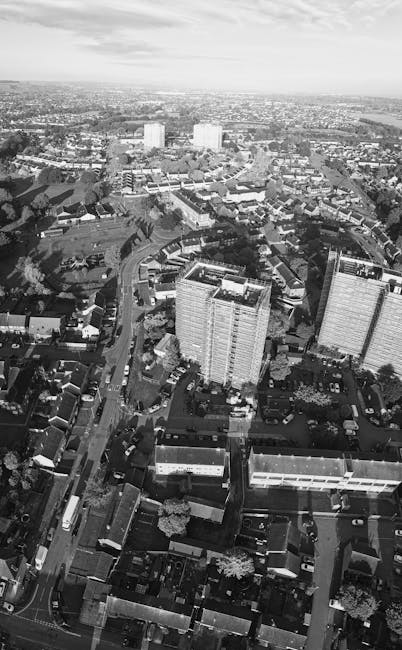 A black-and-white aerial photograph showing a residential area with houses, trees, and a multi-lane motorway running through the scene. The motorway is populated with various vehicles including cars and lorries, and is situated near a suburban neighbourhood with individual houses, gardens, and small streets. In the background, there are open fields and a river or large water body, with a cloudy sky overhead. This image exemplifies a typical urban and rural landscape, often involved in house removals and furniture transport logistics. The scene highlights the transitional environment encountered during home relocation processes, where transportation infrastructure such as busy roads and residential areas intersect with open countryside.